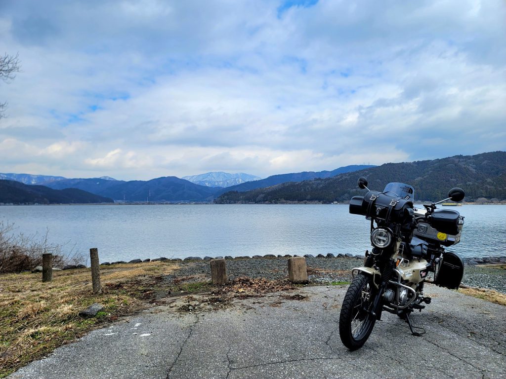 呉湖と遠くの雪山を背景に停まるハンターカブ。湖と山の景色が広がるツーリング風景。