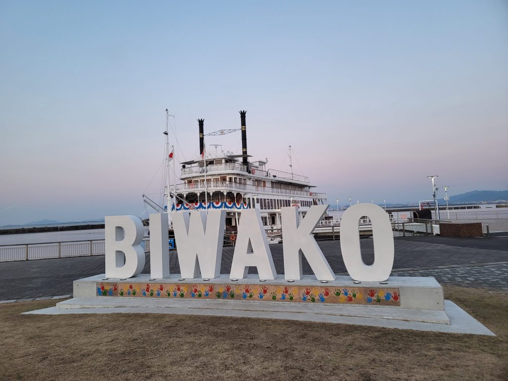 夕暮れの琵琶湖で、BIWAKOモニュメントの後ろにフェリーが映る風景。