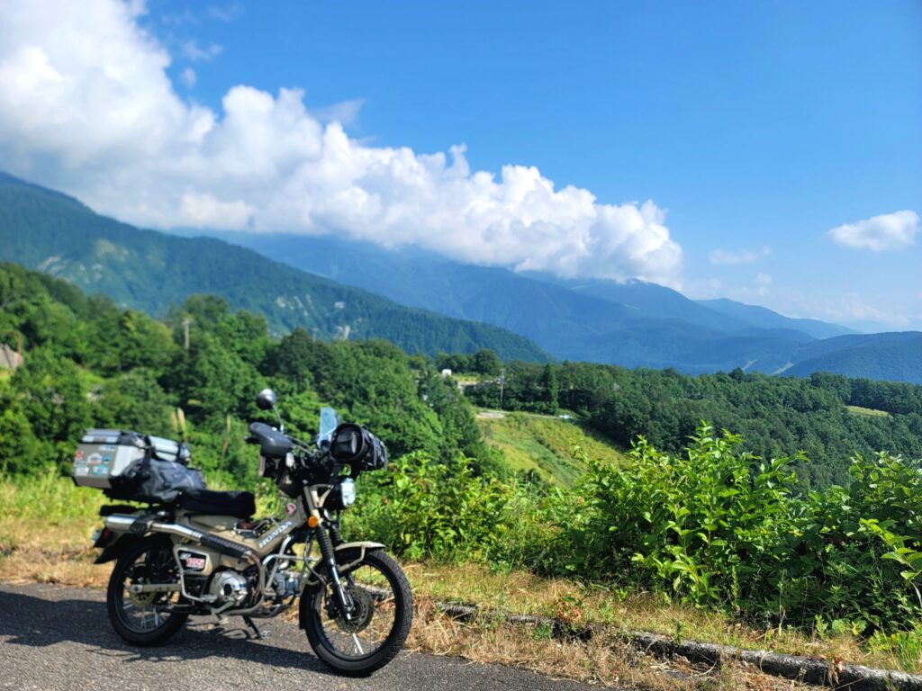 黒菱林道の開けた場所で停めたハンターカブと、横に広がる山々、近くの空と雲が映る林道ツーリングの写真。