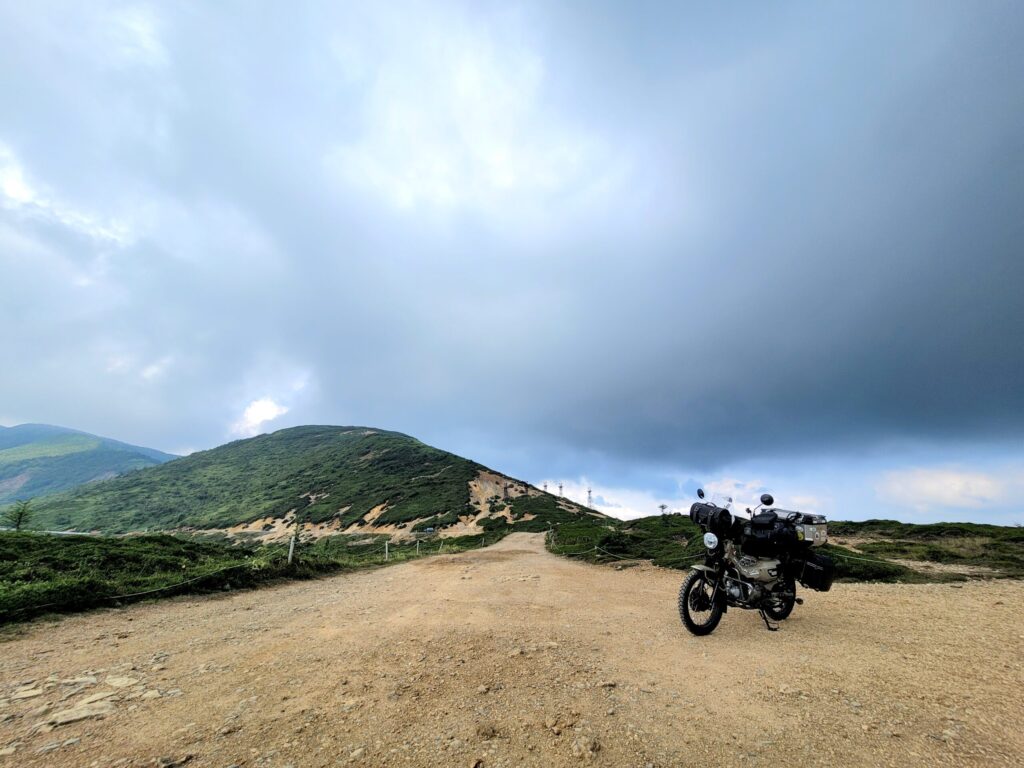 毛無峠のダート林道に停めたハンターカブの写真。足元の路面や遠くの山々、送電線も見える峠の様子。