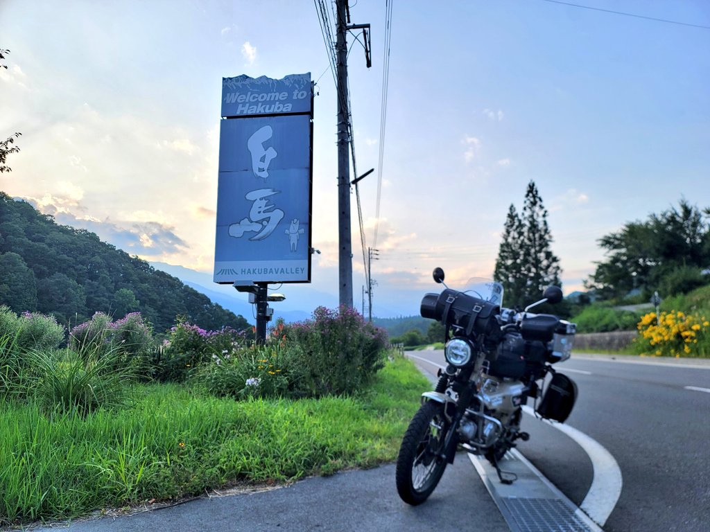 白馬村の看板前でハンターカブと記念撮影。夕暮れで少し薄暗く、柔らかい光に包まれた景色。