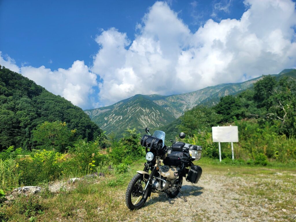 白馬エコーランドの平川沿いに停めたハンターカブと、背景に広がる北アルプスの山々。高原の爽やかな景色とバイク旅の雰囲気が伝わる写真。