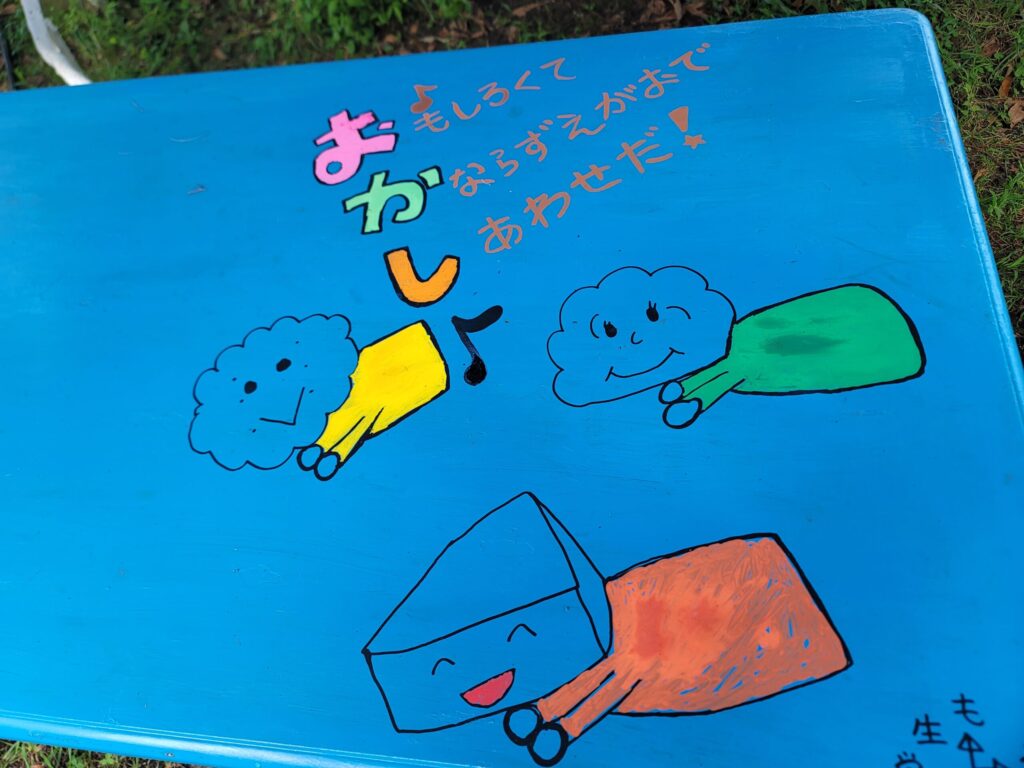 長野、おかしの家くるみぼーるの外のテーブルに描かれたかわいいイラスト。空を飛ぶキャラクターと「かならずえかおで、しあわせだ」と文字がデザインされている。