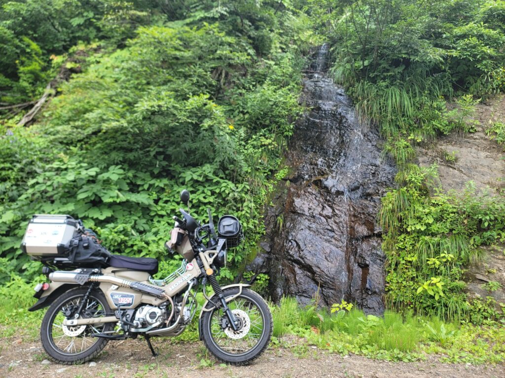 妙高小谷線、林道途中の小さな滝の前に停めたハンターカブの写真。長野県側は緩やかな登りで走りやすい様子。