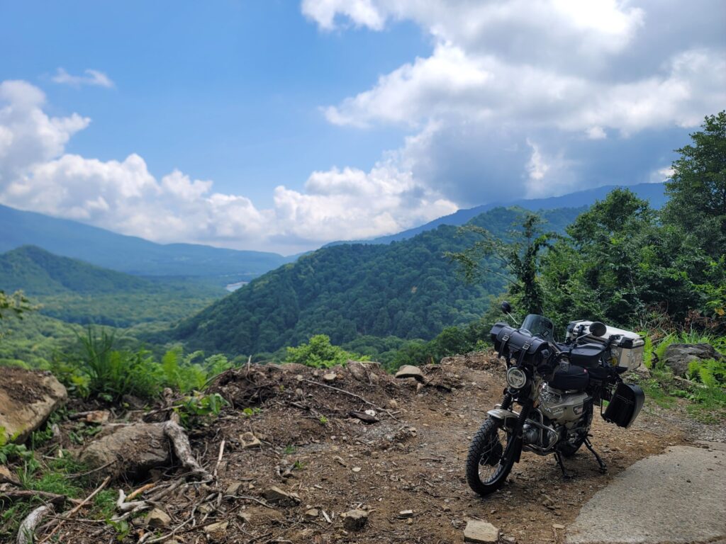 妙高小谷線、林道の絶景ポイントでハンターカブを停め、背景に広がる山々と空を撮影した写真。