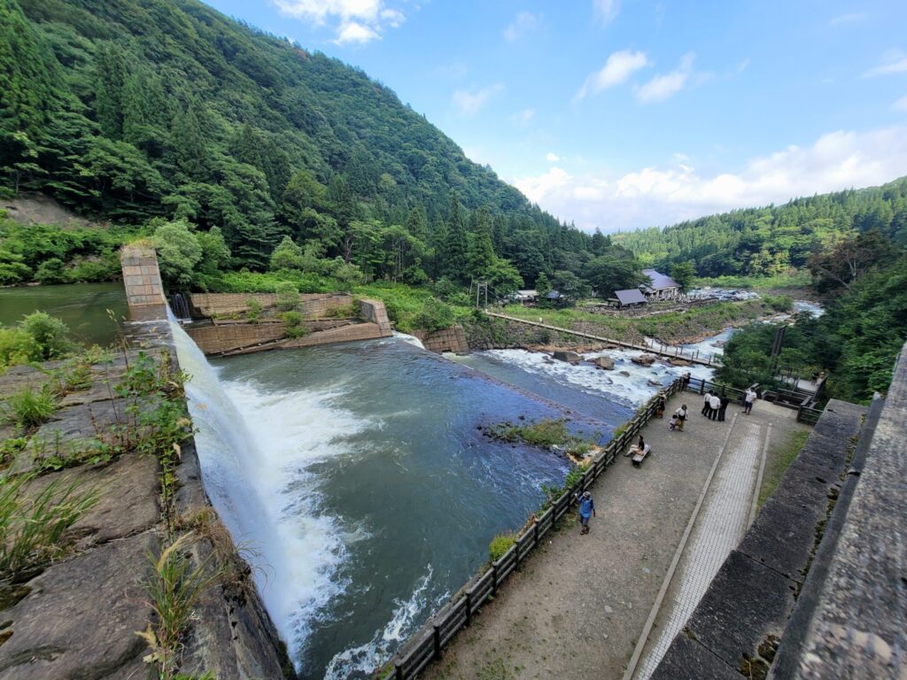 新潟、苗名滝の堰堤から勢いよく流れ落ちる水の様子。下流にはカフェと吊り橋が映り込む、自然と観光スポットが一体となった景観。