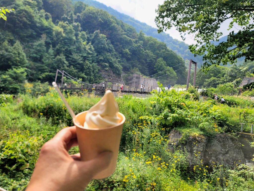 新潟、苗名滝そばのCafe Glandで、砂防堰堤を背景にソフトクリームを持つ写真。バイクツーリング途中の休憩風景。