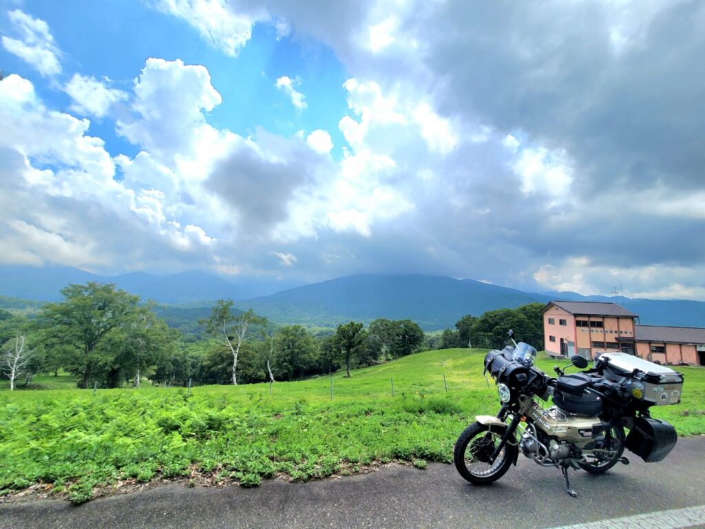 新潟、笹ヶ峰牧場の緑の草原の中で、牧舎の前に停めたハンターカブと記念撮影している写真。