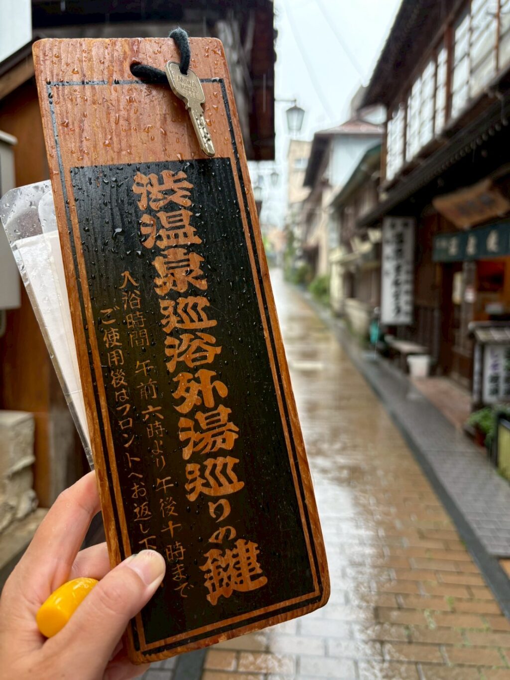 長野・渋温泉の外湯めぐりで使う木札の鍵を接写した写真。背景に温泉街の町並みがぼんやり写っている。バイクツーリングの疲れも外湯で解消。