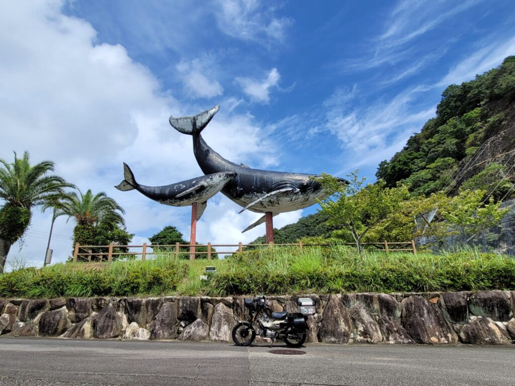 和歌山県太地町の実物大の親子クジラのオブジェと、横に停めたハンターカブ。青空を背景にクジラが泳いでいるように見える風景。