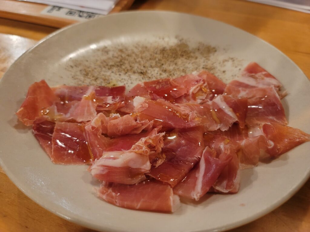 長野・白馬のイタリアンレストラン「Tomatito」で提供されるスペイン産の原木生ハムの写真。大きな白いお皿に生ハムが半分ほど盛られ、もう半分には粒胡椒が散らされた美しい盛り付け