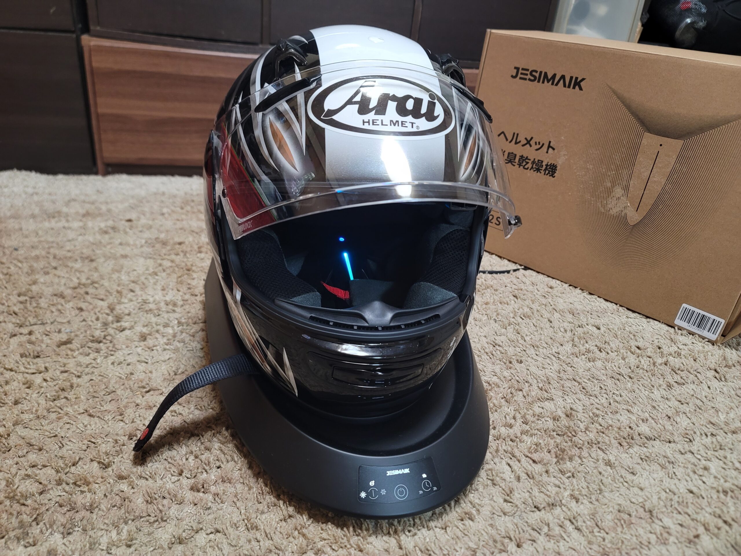 JESIMIAKヘルメット消臭乾燥機A2Sの上に息子のヘルメットを置き、電源を入れた状態。 稼働中のLEDランプが青・緑・赤と順に光り、除菌と乾燥が進んでいる様子。